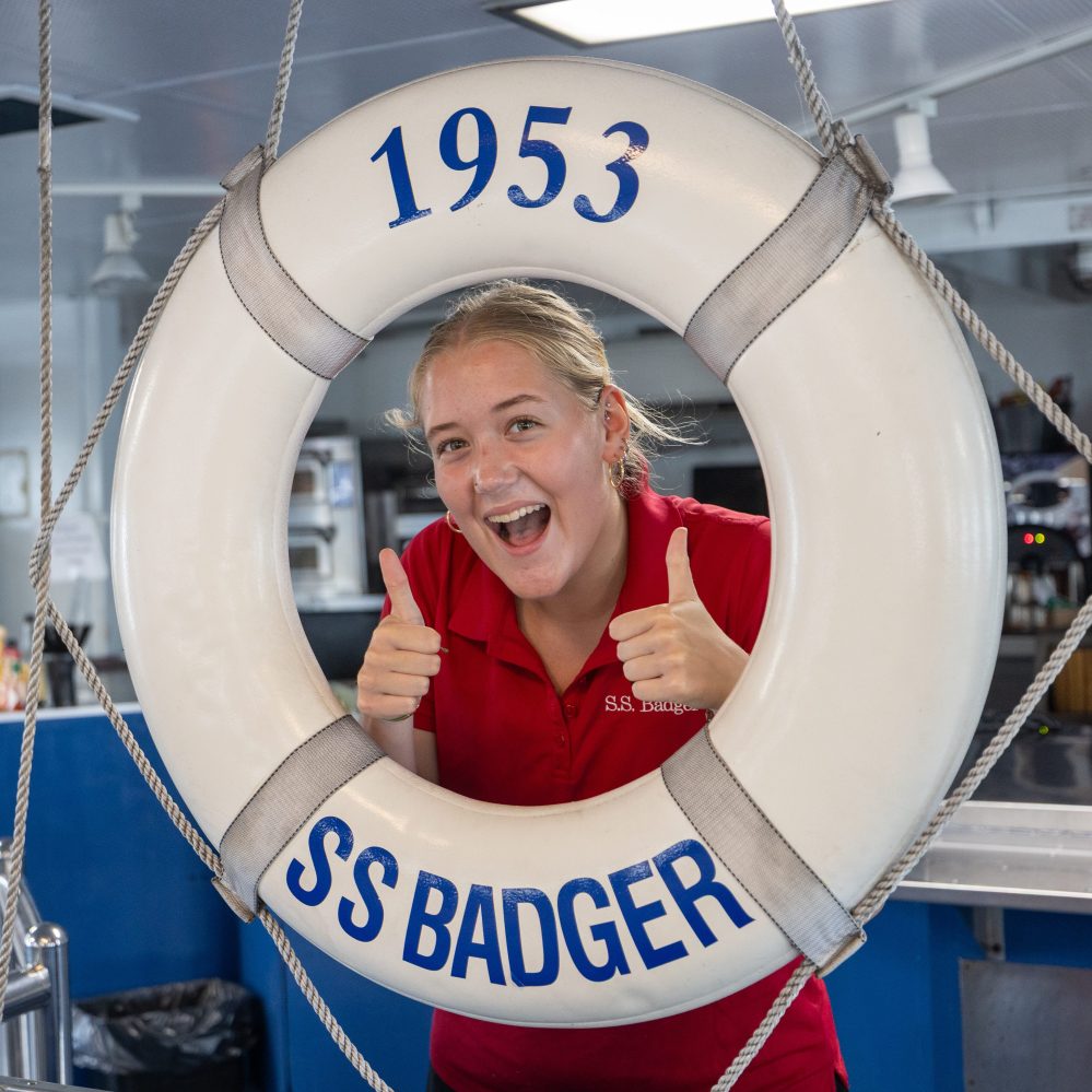 Person smiling with two thumbs up through SS Badger life ring on a boat.