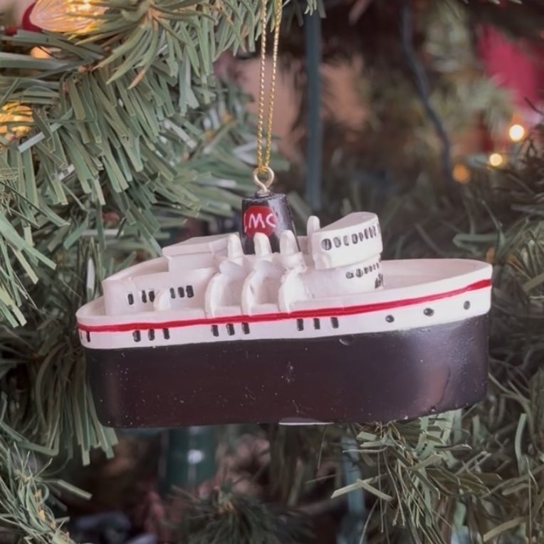 Ship ornament hanging on a Christmas tree with lights and green branches.