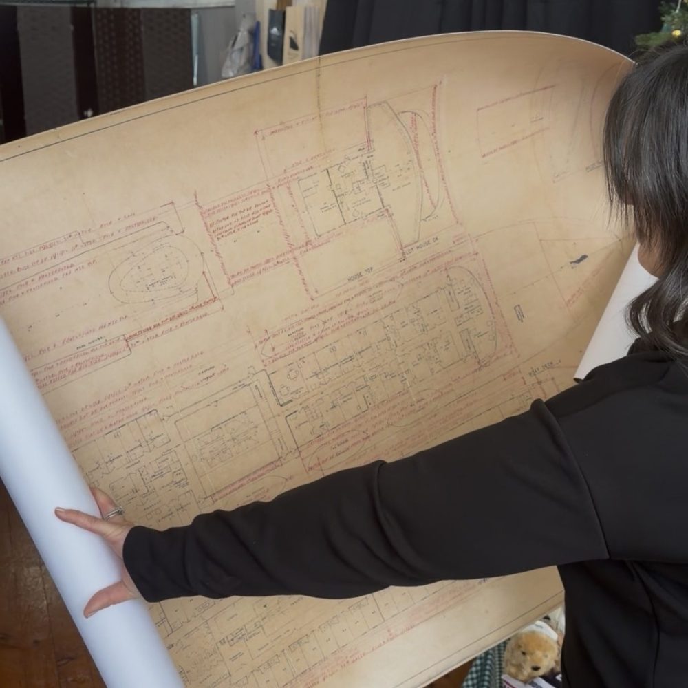Person holding a large blueprint with architectural drawings in a room with wood floor and dark curtains.