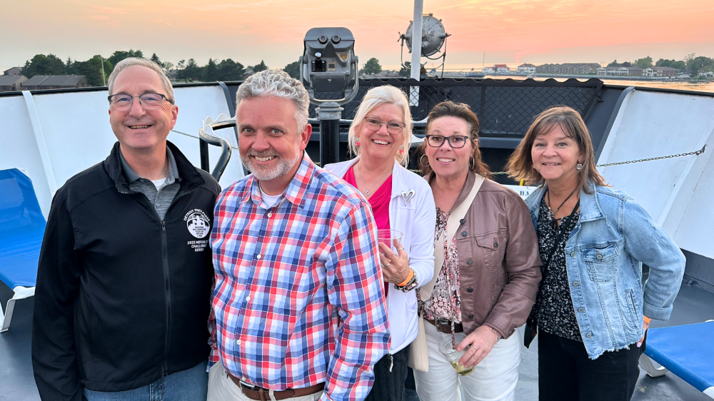 Five people posing on a boat at sunset, smiling and standing close together.