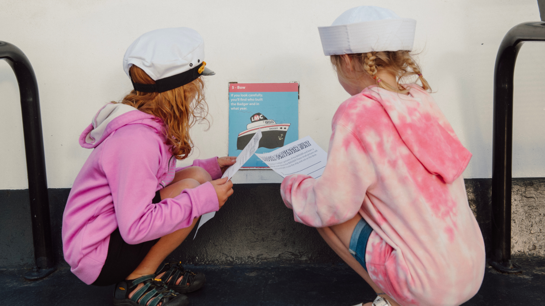 Two children in sailor hats read a sign on a boat wall.