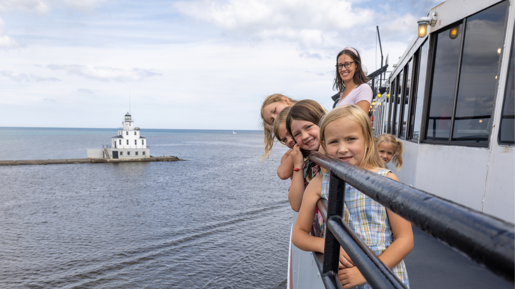 Children and a woman on a boat near a distant lighthouse over water.