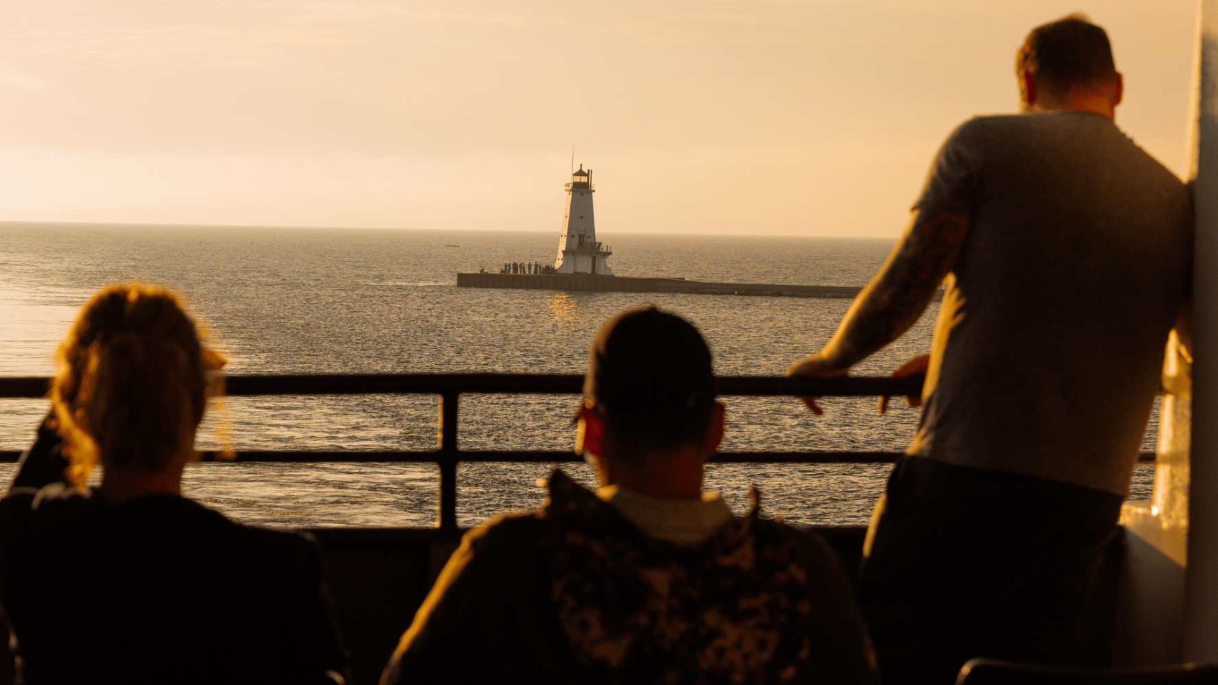 Three people on a boat, overlooking a lighthouse on the ocean at sunset.