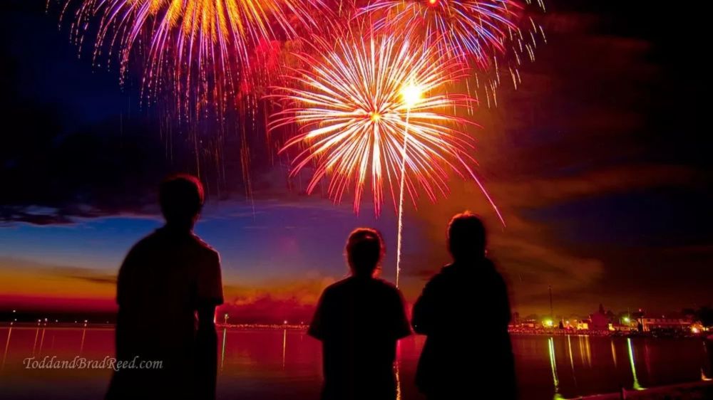 Silhouetted people watching vibrant fireworks over water at dusk.