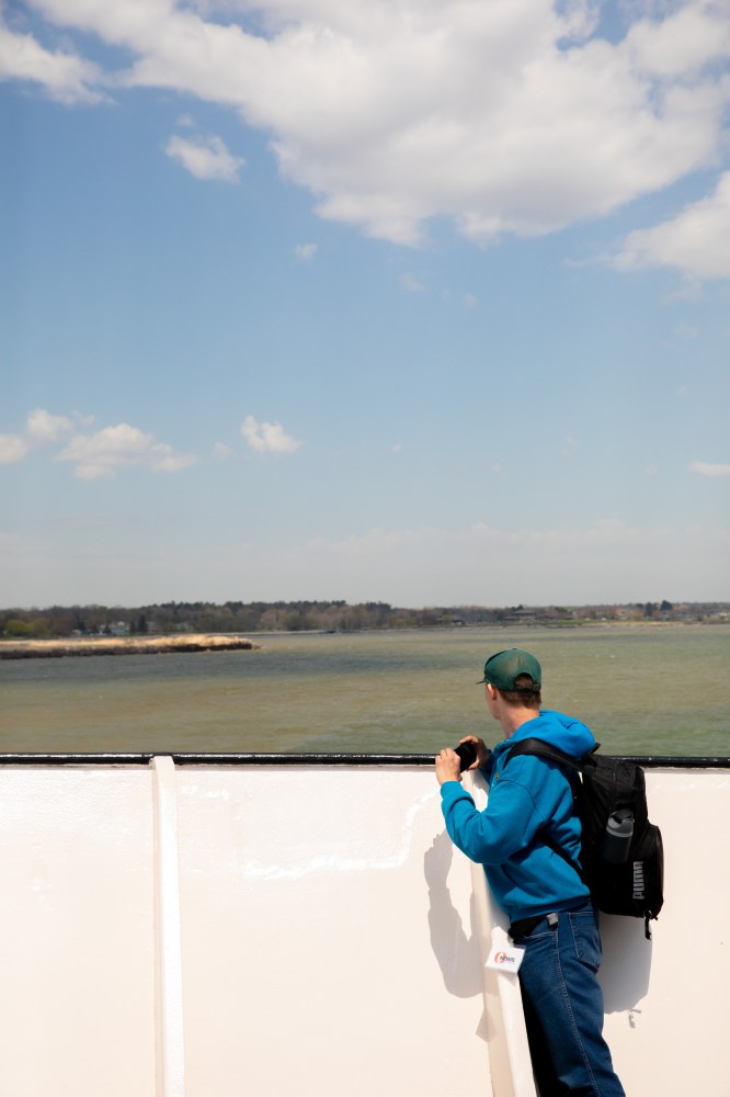Person in blue hoodie on ferry looks at distant shoreline under a cloudy sky.