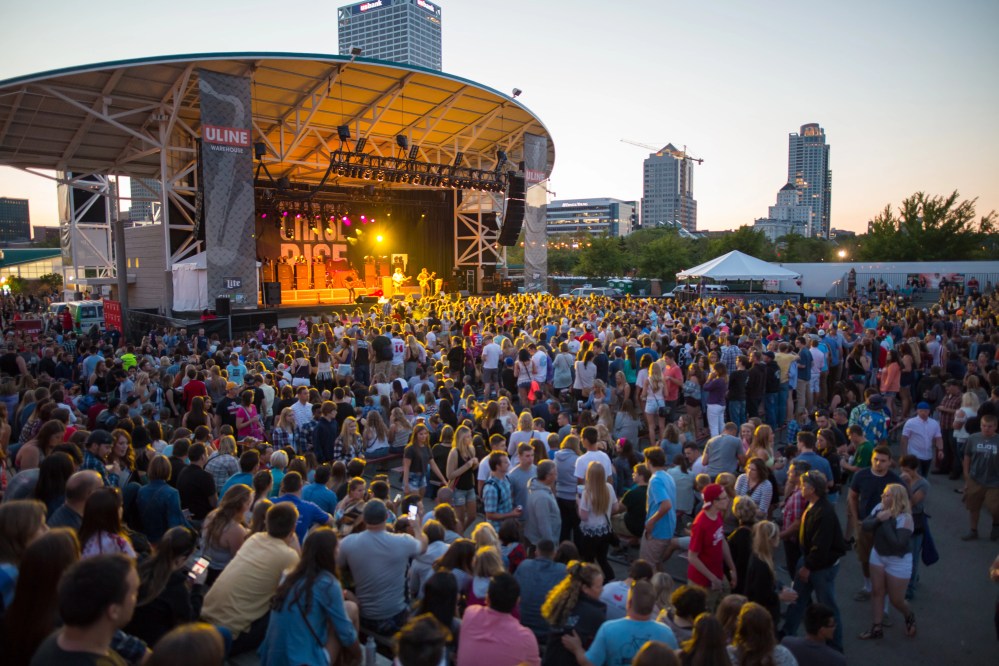 Crowd at outdoor concert venue with city skyline in background at sunset.