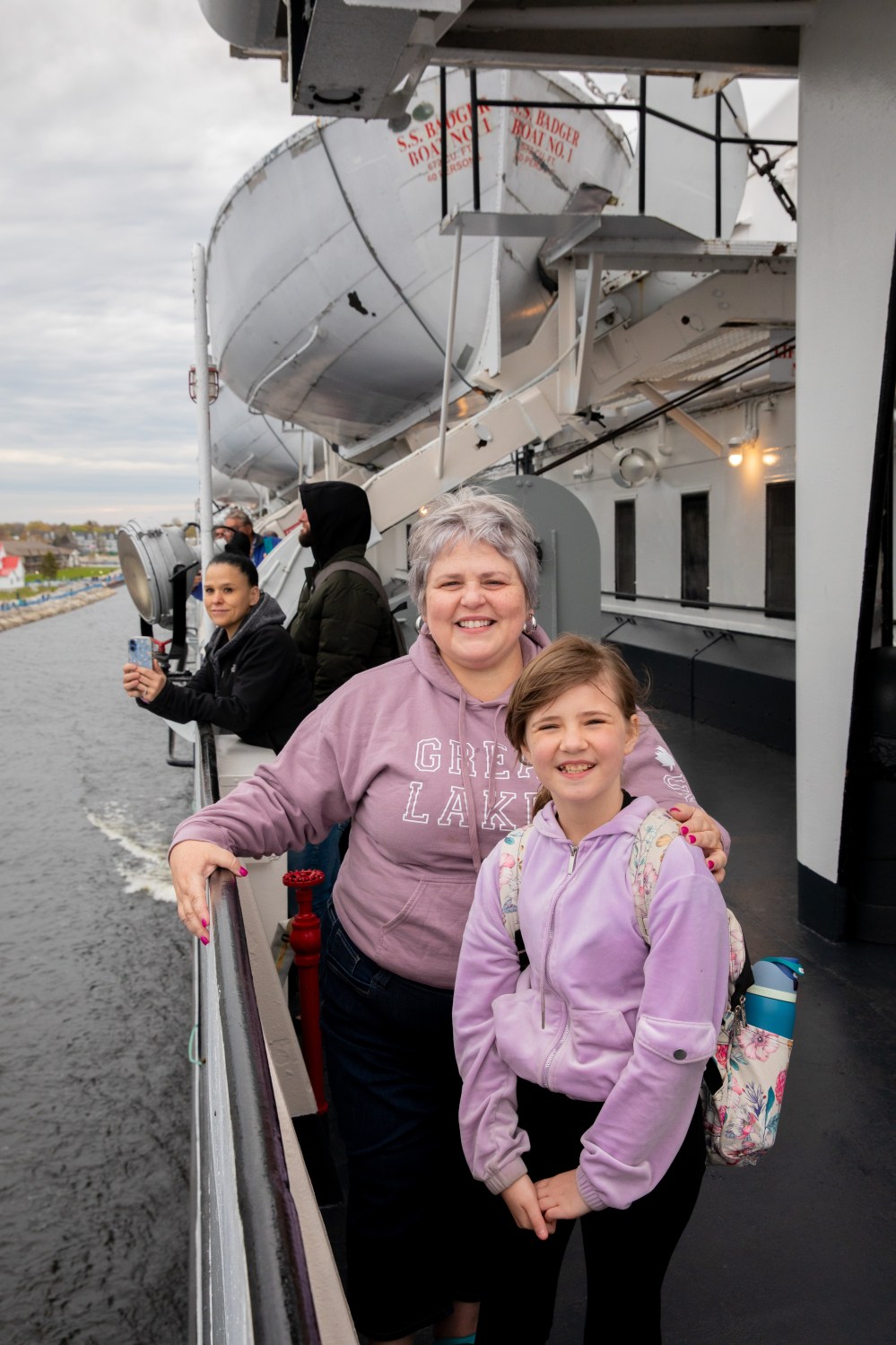 Smiling woman and girl on a ferry deck with others and a lifeboat in the background.