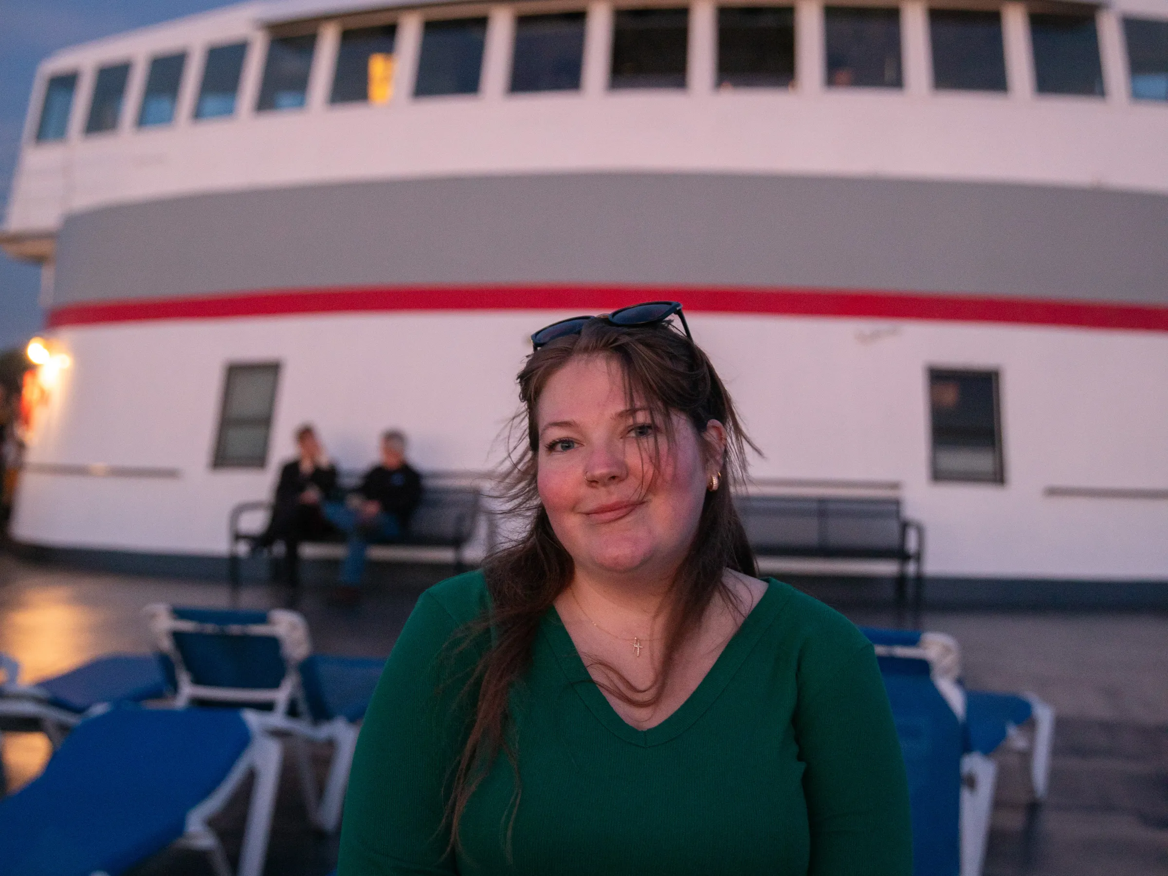 Person in green shirt stands on boat deck at dusk, with ship's bridge in background.