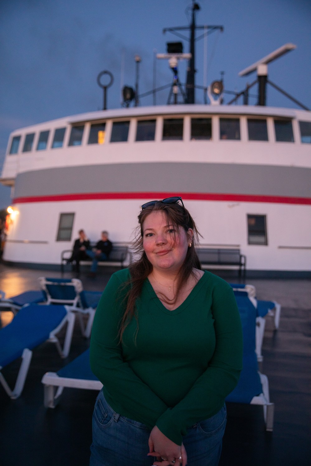 Person in green shirt stands on boat deck at dusk, with ship's bridge in background.