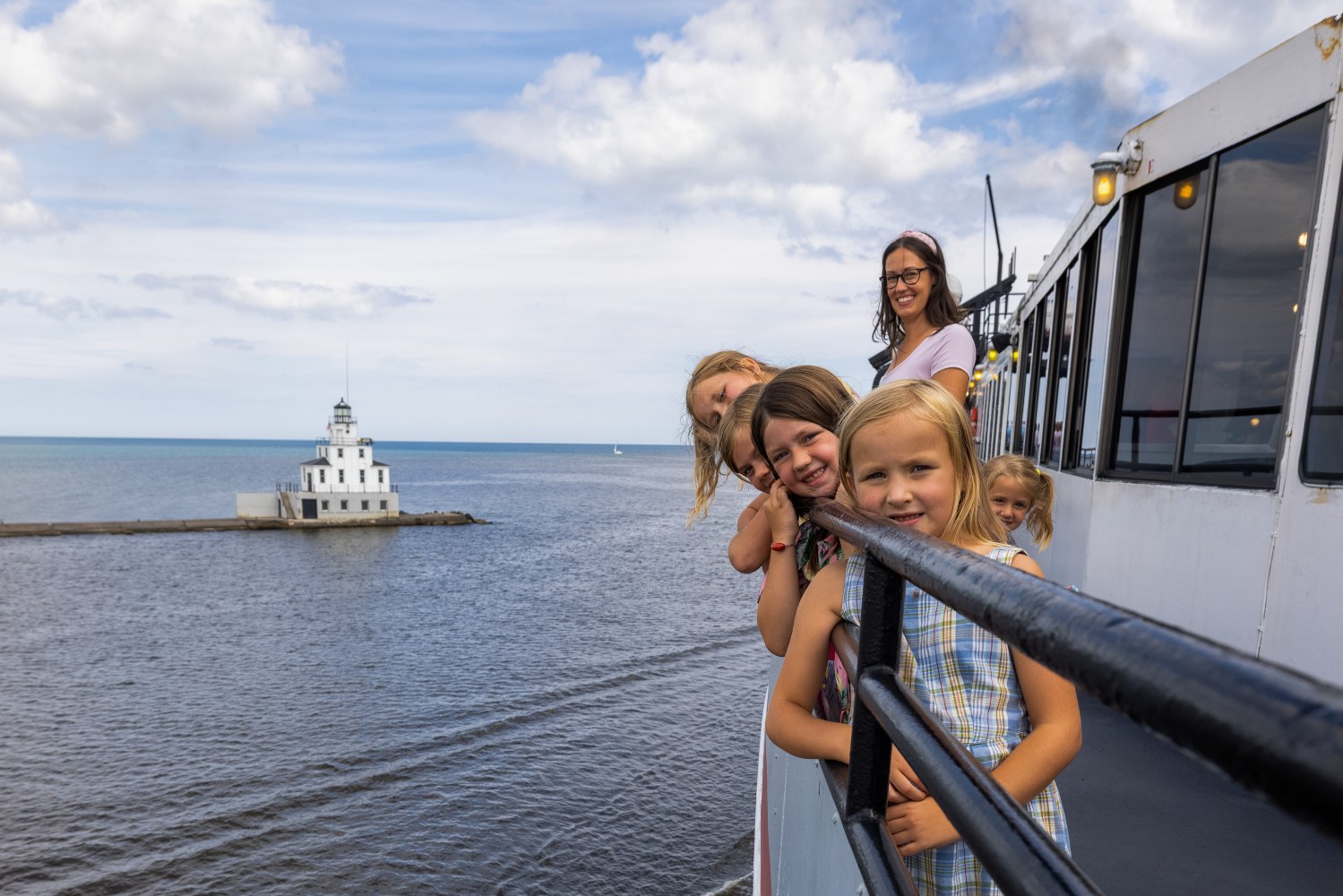 Group of children on a boat smiling at camera, lighthouse in background.