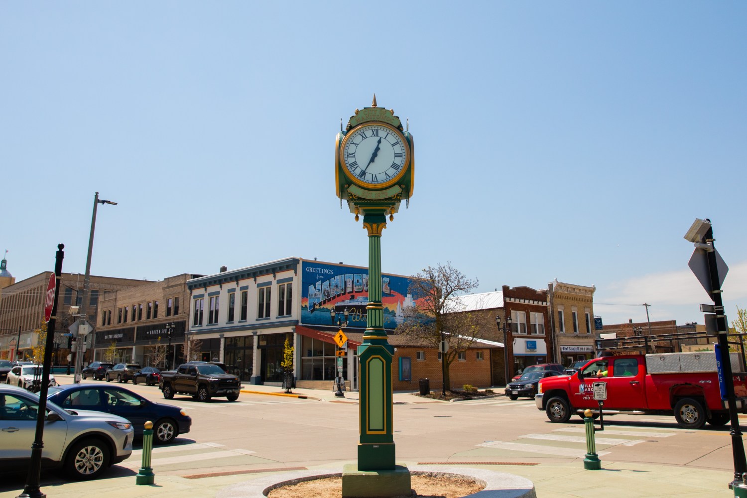 Street with vintage clock, cars, and buildings under clear sky.