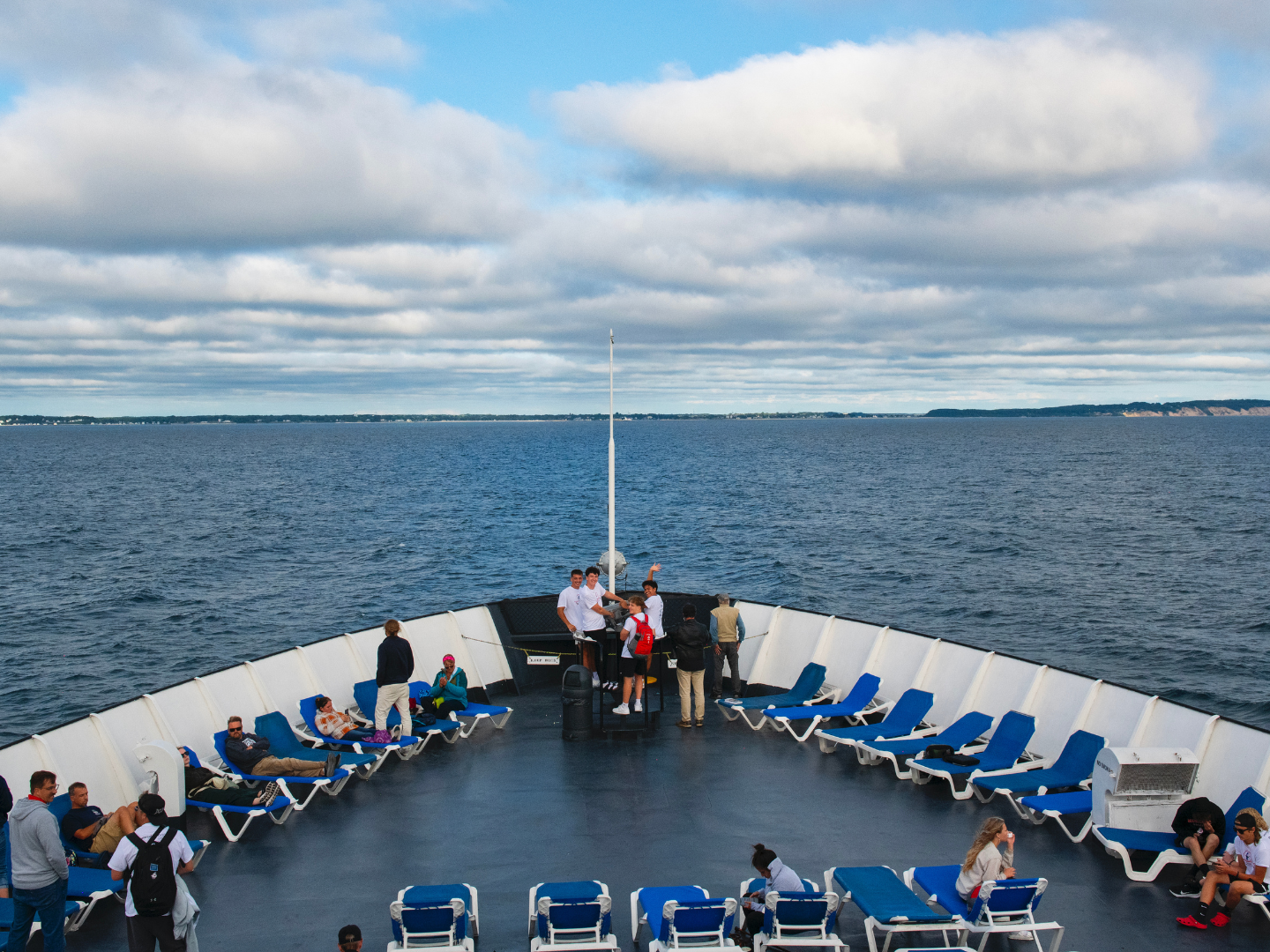 People on a ship deck with blue lounge chairs overlooking the sea.