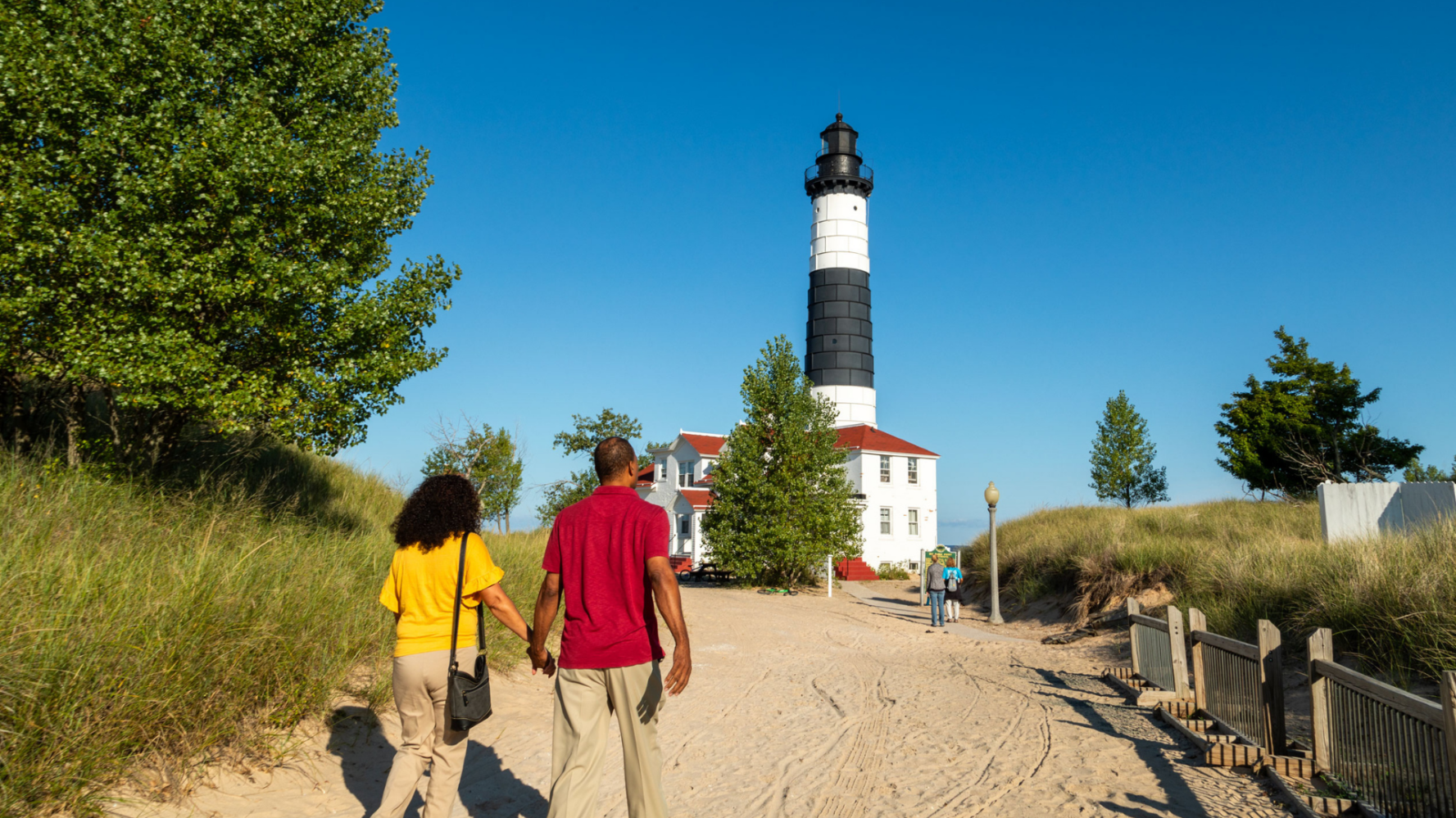 Two people walk towards a lighthouse on a sandy path.