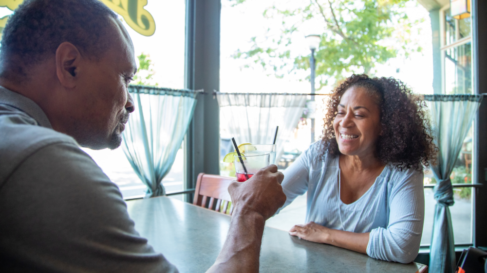 Two people smiling, sitting at a cafe table with drinks by a window.